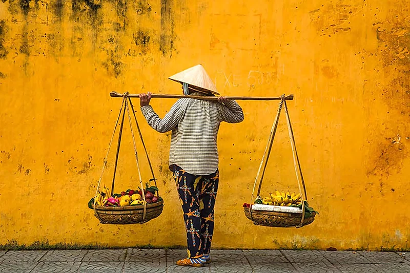 Vendedora con canastos de fruta al hombro frente a una pared amarilla en Hoi An