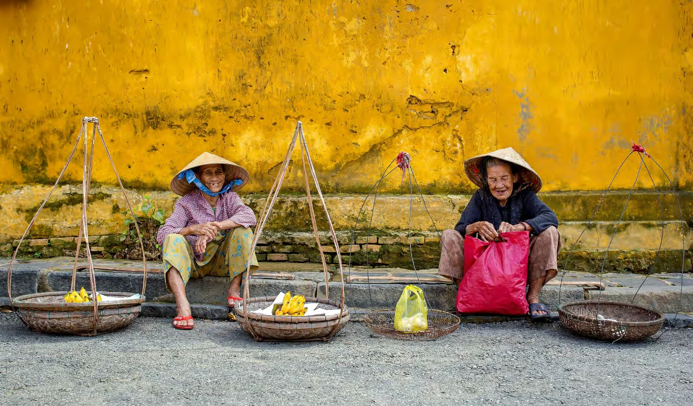 Dos vendedoras con sombrero cónico y sus canastos frente a una pared amarilla en Hoi An