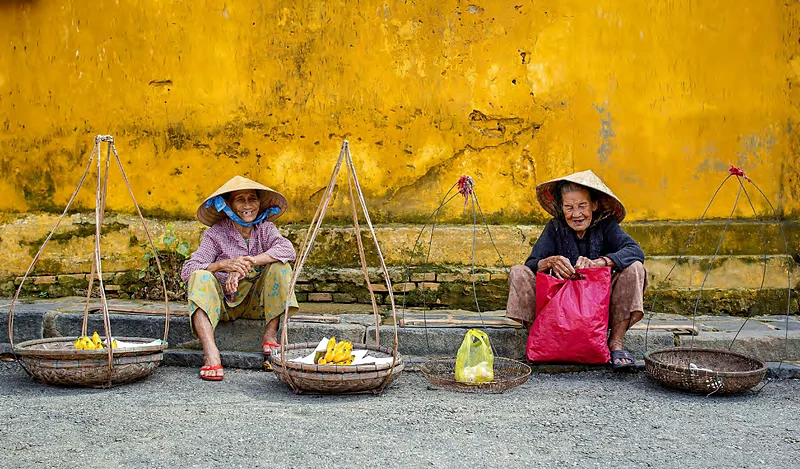 Dos vendedoras con sombrero cónico frente a una pared amarilla en Hoi An